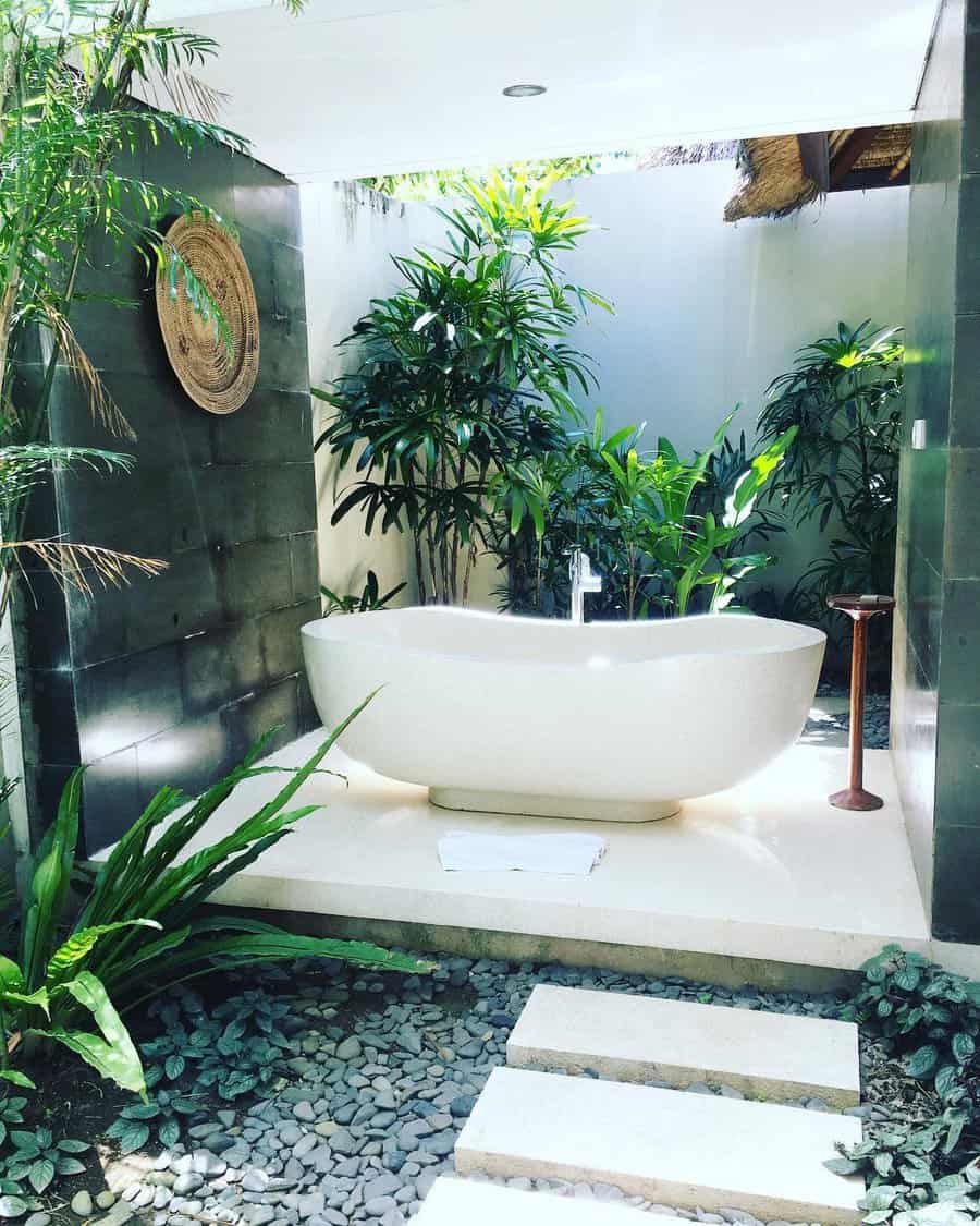 Outdoor bathroom with a white bathtub surrounded by lush green plants, stone walls, and a pebble pathway