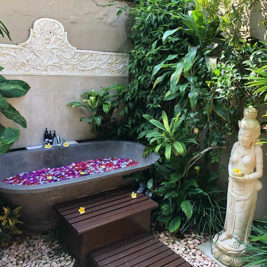 Outdoor stone bathtub with floating flower petals, surrounded by lush greenery, a wooden mat, and a statue of a serene figure