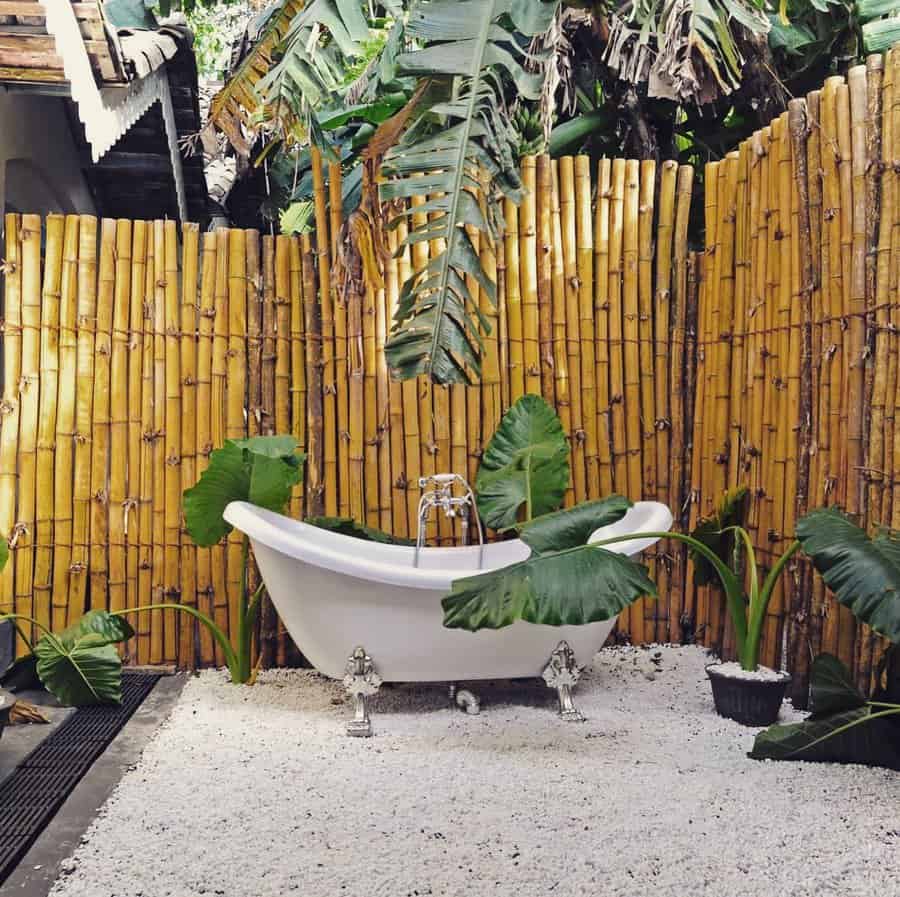 Outdoor bathtub surrounded by bamboo fencing and tropical plants, set on a gravel surface under lush greenery