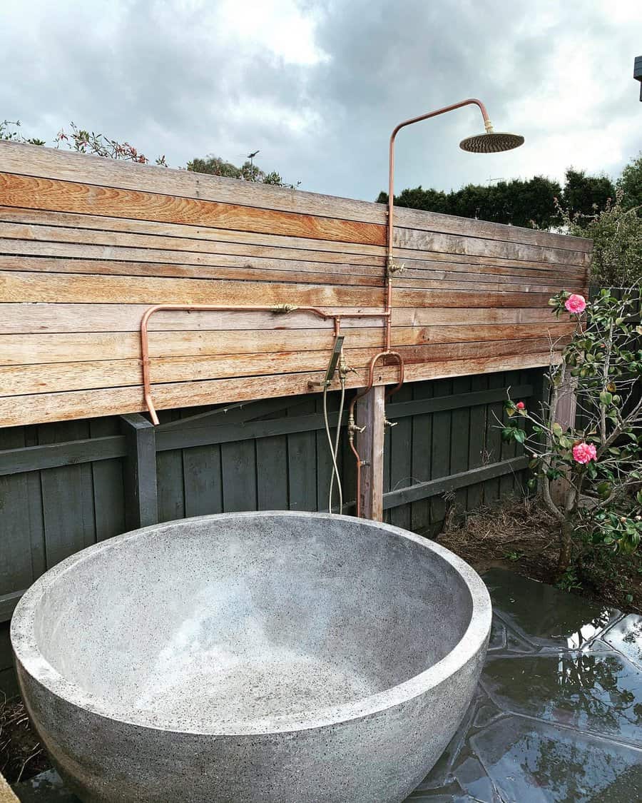 Outdoor round stone tub with copper shower fixtures, wood fence, and flowering plant on a cloudy day