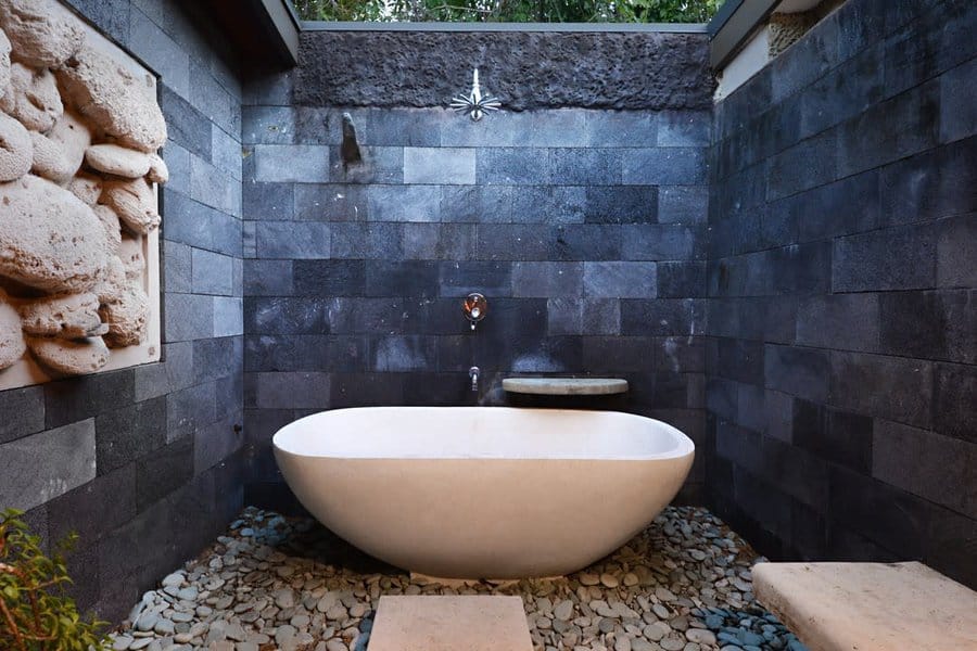 Outdoor bathroom with stone walls, featuring a white freestanding bathtub on a pebble floor and a small wall-mounted shelf