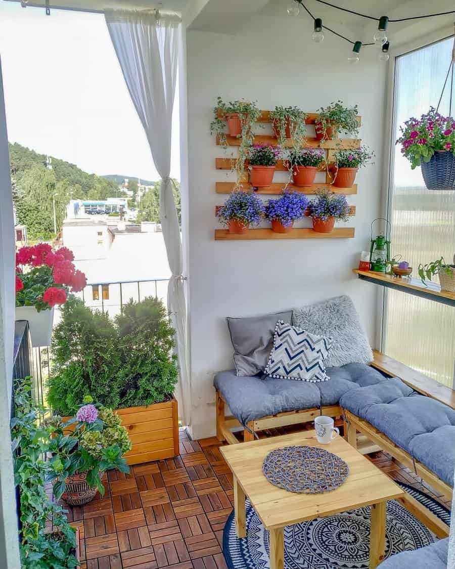 Small balcony with a cushioned seating area, coffee table, plants on shelves, hanging plants, and a view of trees and buildings