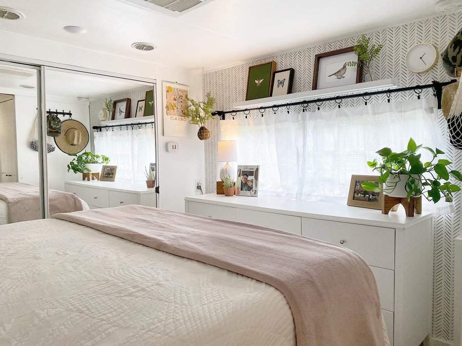 Bright RV bedroom with white dressers, mirrored closet doors, and decorative plants, creating a spacious and organized retreat.