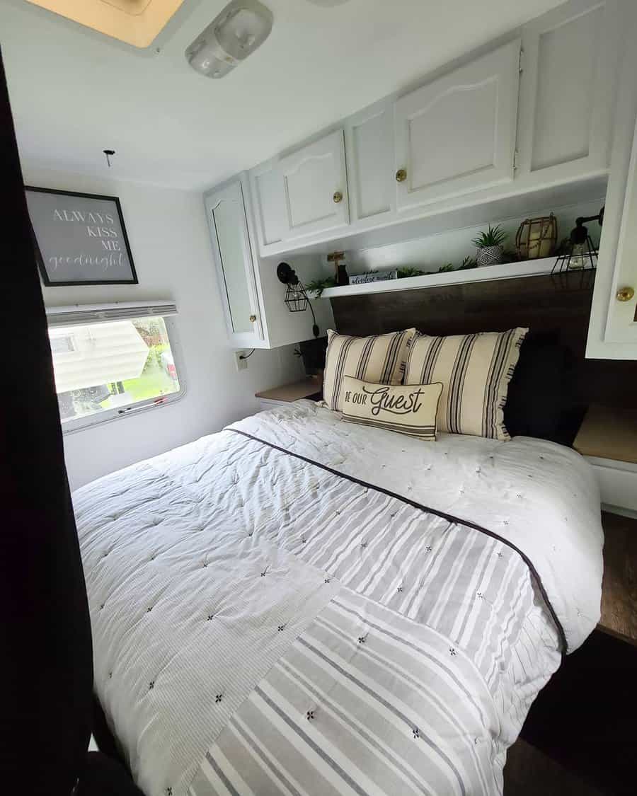 Cozy RV bedroom with overhead white cabinets, a built-in shelf, and striped bedding, blending storage with a welcoming design.