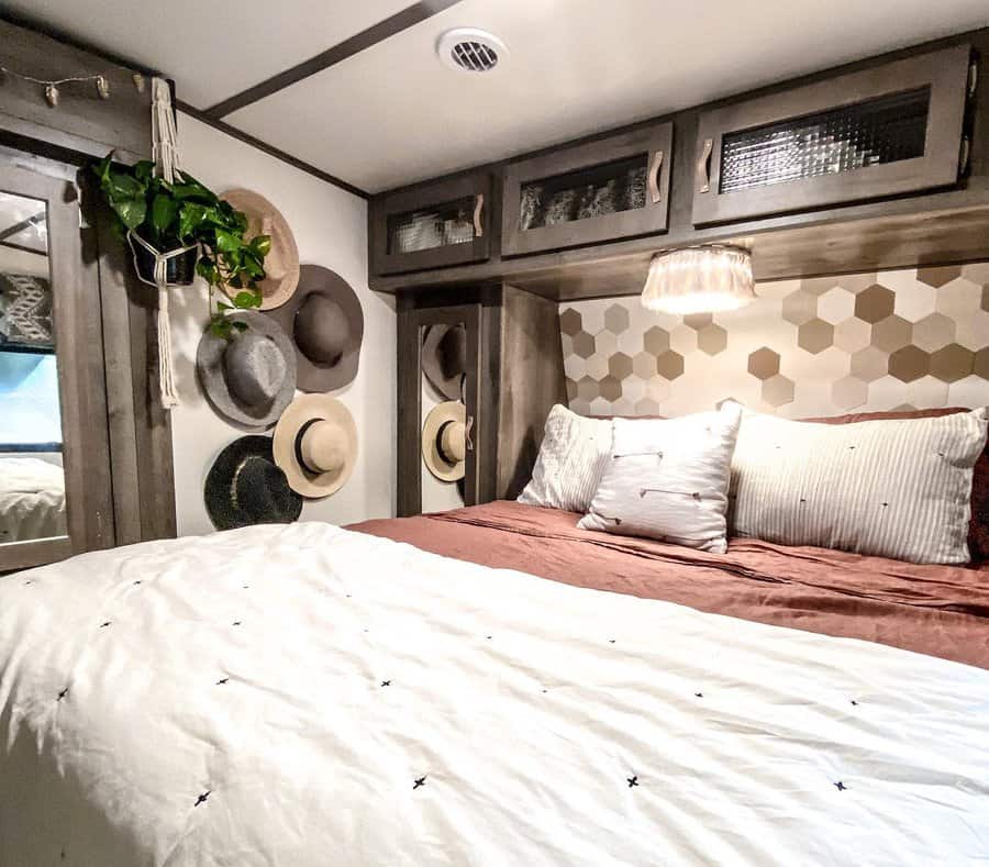 Rustic RV bedroom with overhead cabinets, a hexagon accent wall, hat decor, and warm bedding, combining storage with cozy charm.