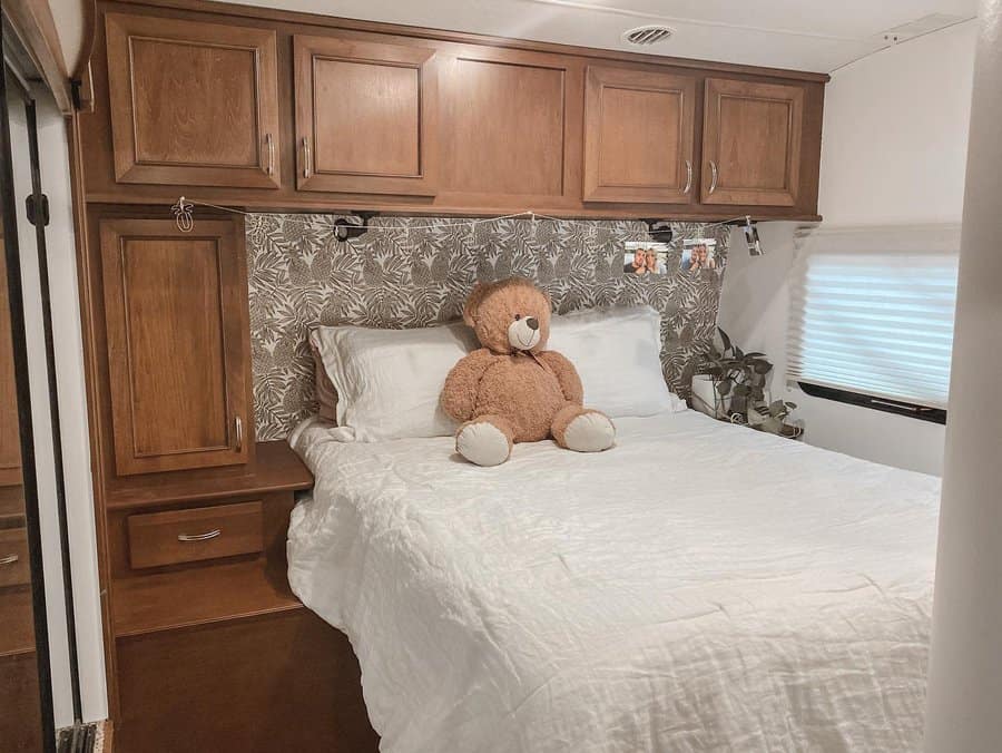 Warm RV bedroom with wooden overhead cabinets, a cozy bed, a teddy bear, and a patterned accent wall, blending comfort with storage.