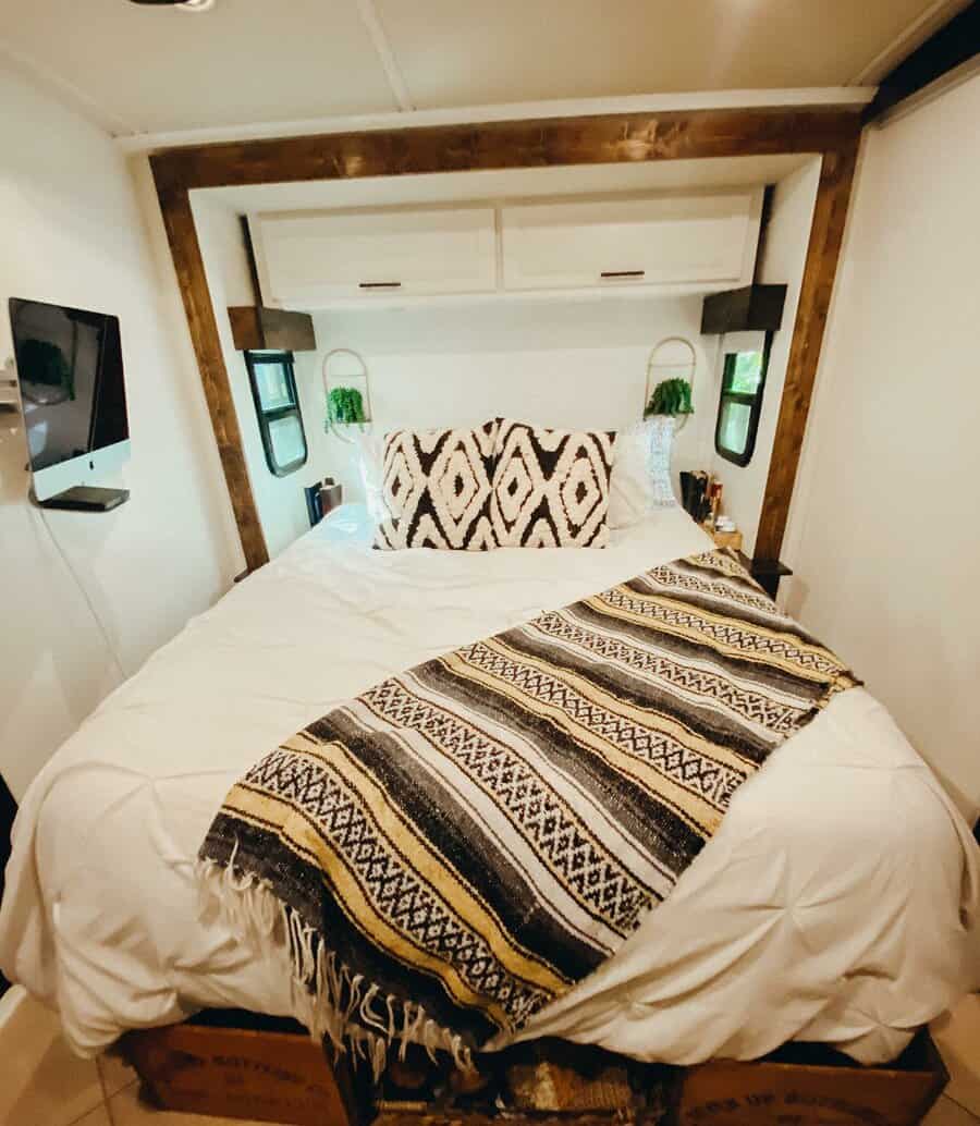 Cozy RV bedroom with a rustic wood frame, white overhead cabinets, geometric pillows, and under-bed storage, blending style and function.