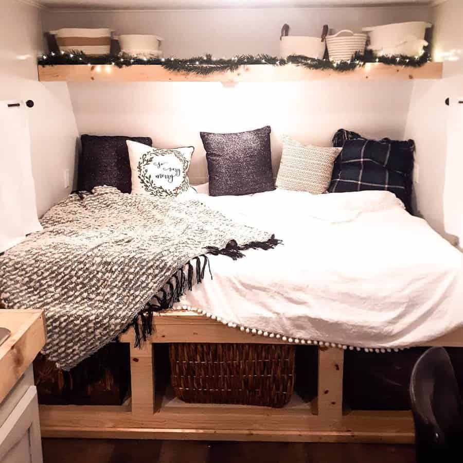 Cozy RV bed with built-in wood storage, decorative pillows, soft blankets, and overhead shelving with baskets and string lights.