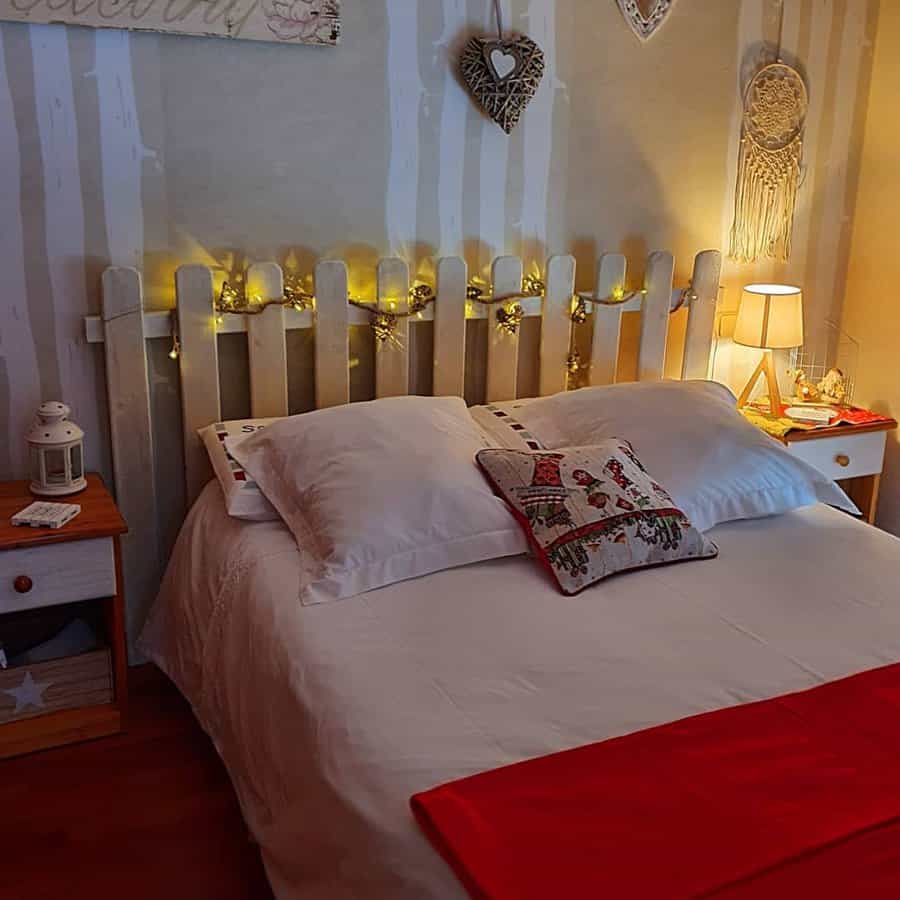 Cozy bedroom with a wooden headboard, fairy lights, dreamcatchers, and a red bed runner. A small lamp and decor items on side tables