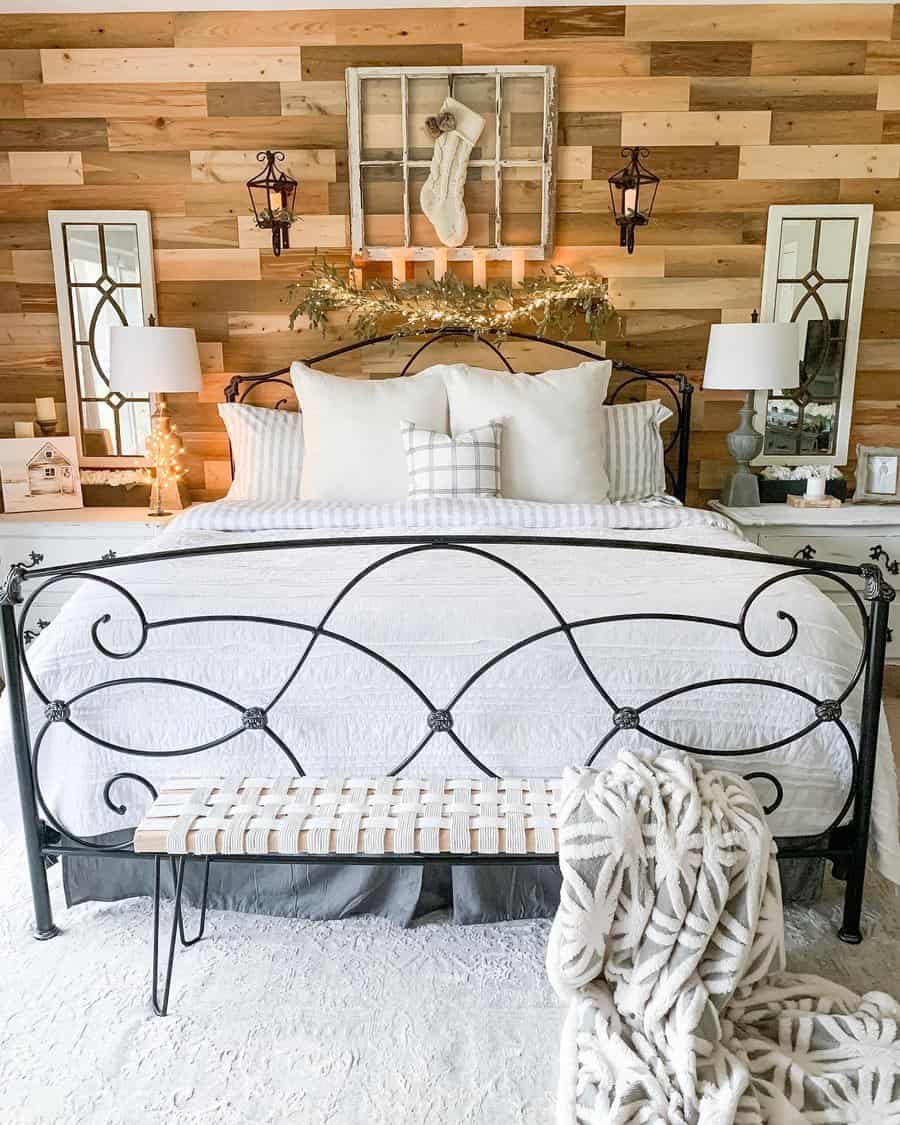 Cozy bedroom with a wrought iron bed, striped bedding, wooden accent wall, and festive garland with string lights, with two lamps and mirrors flanking the bed