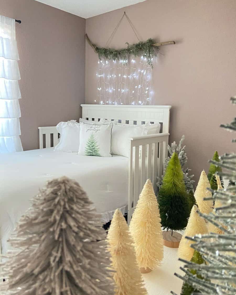 A cozy bedroom with a white bed, pine-themed pillows, and a decorative branch with string lights, mini Christmas trees line the front