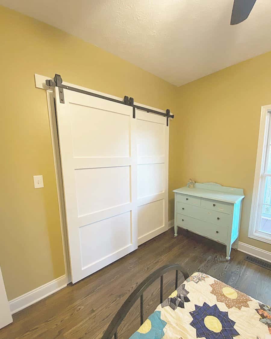 White barn-style sliding closet doors with black hardware add a rustic touch to a cozy bedroom with warm yellow walls and wood flooring