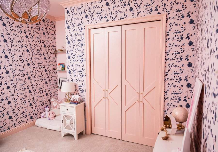 A room with floral wallpaper, light pink closet doors, a white nightstand with a lamp, and stuffed toys on a cushion