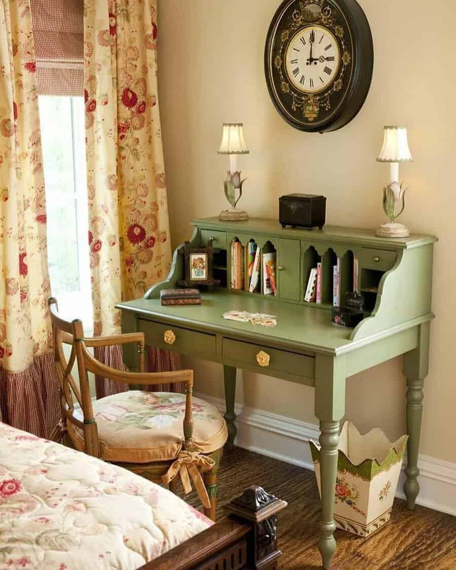 A vintage green desk with books and lamps, a floral chair, a flowered curtain, and a wall clock above in a cozy room setting