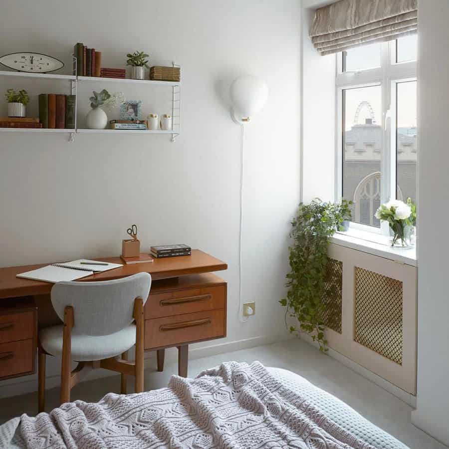 Cozy bedroom with a wooden desk and chair, wall shelves with books, a bed with a knit blanket, and a window with plants and a city view