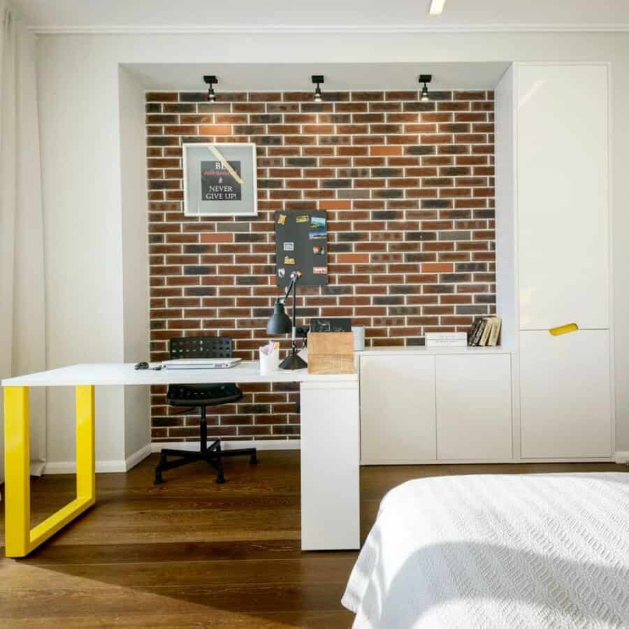 Home office with a desk, chair, and lamp against a brick accent wall. Mounted artwork reads "NEVER GIVE UP" Cabinets and a bed are visible