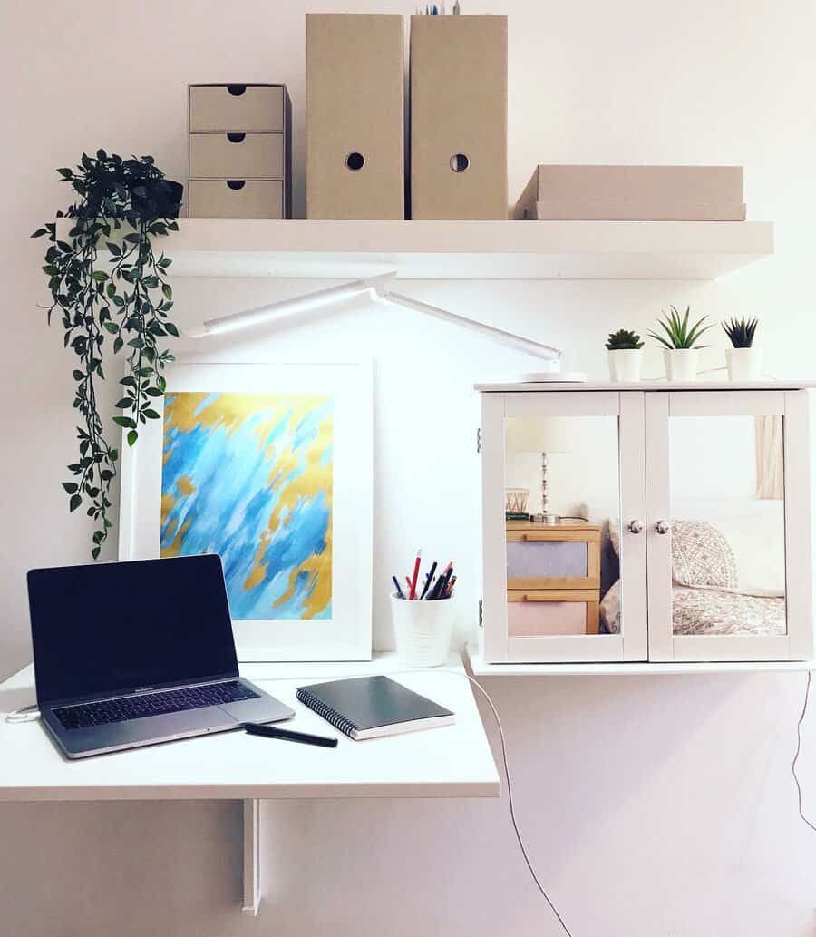 Minimalist workspace with a laptop, notebook, wall art, and mirror cabinet on a white desk; shelves hold plants and storage boxes