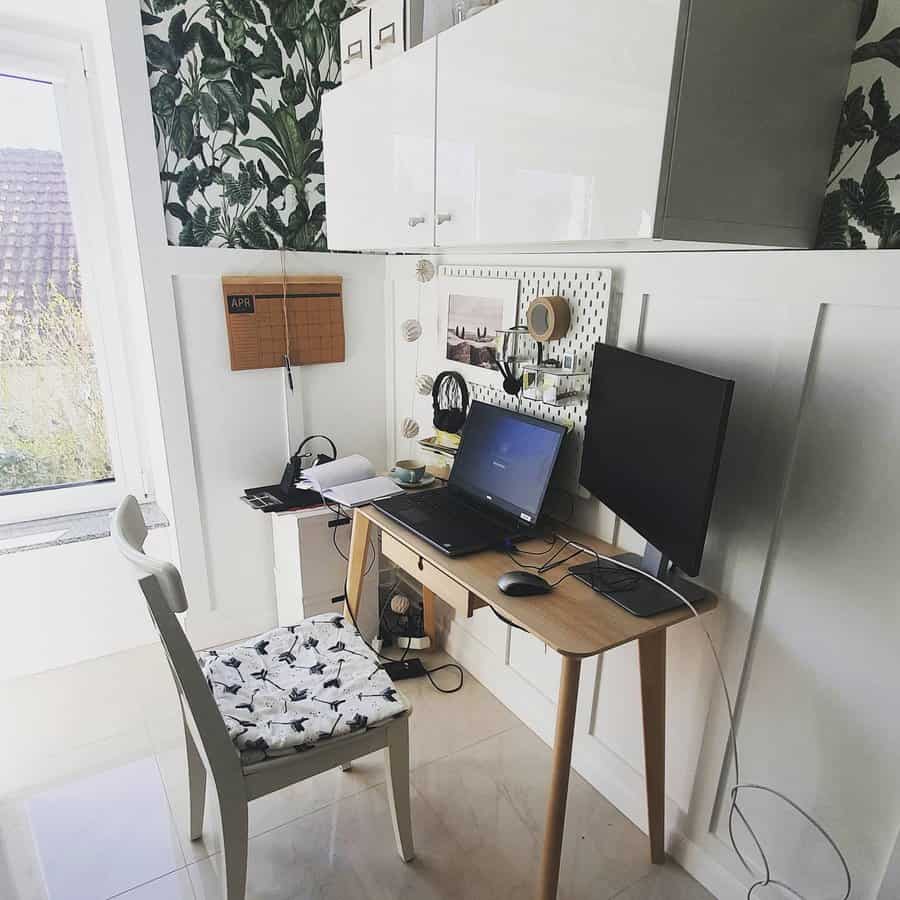 Compact home office with a wooden desk, laptop, monitor, and chair, green leaf wallpaper and window with a garden view