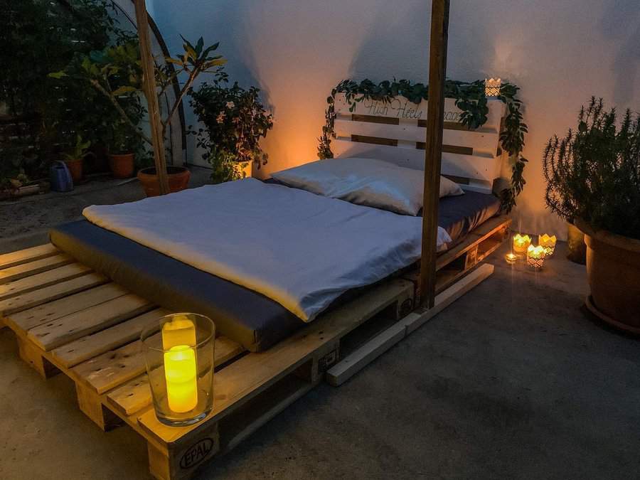 Outdoor bed on a pallet platform with cozy bedding, surrounded by plants and lit candles, creating a warm, inviting atmosphere