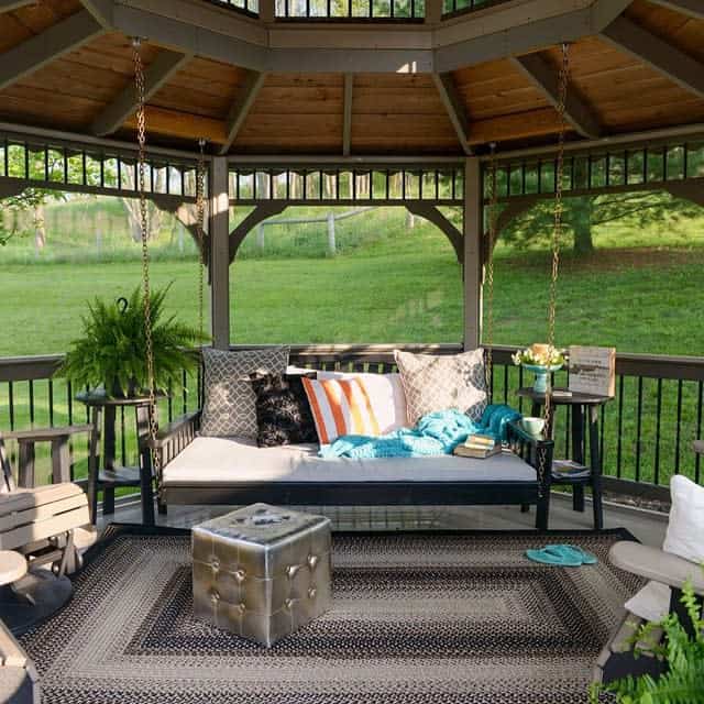 A cozy gazebo with a hanging bench, cushions, a blue throw, and a small table, surrounded by greenery on a sunny day