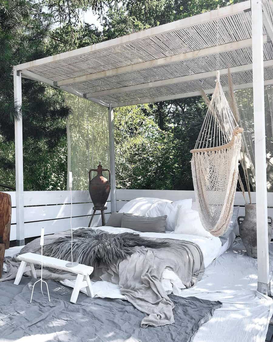 Outdoor canopy bed with white pillows and gray throw, surrounded by greenery. A hammock chair hangs nearby; rustic, cozy setting