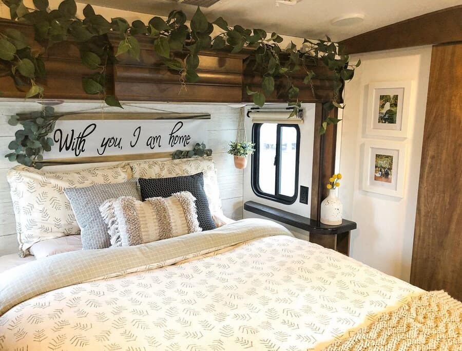 A cozy RV bedroom with soft, neutral bedding, a framed sign, hanging plants, and framed artwork, creating a homey and inviting atmosphere.