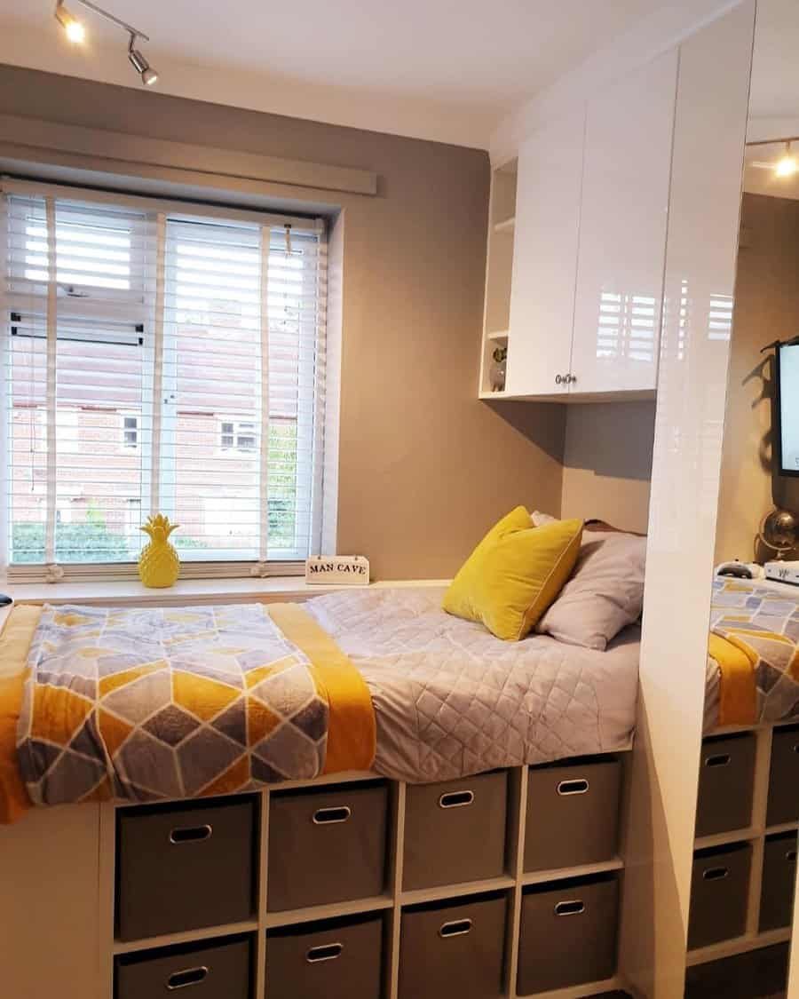 A space-saving bedroom with a raised bed featuring built-in cube storage, overhead cabinets, and a cozy yellow and gray color scheme