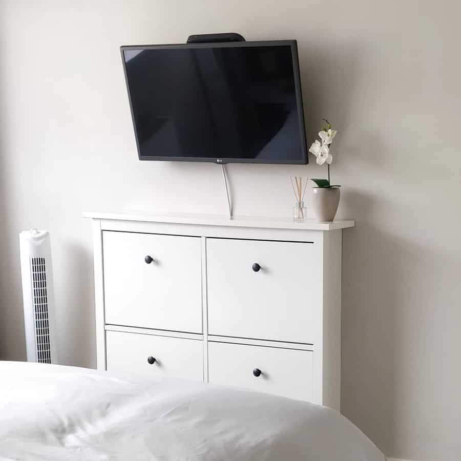 Wall-mounted TV above a white dresser with four drawers, a small plant, and reed diffuser in a minimalist room