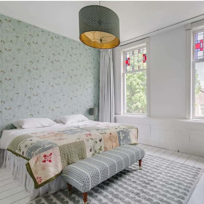 A cozy bedroom with floral wallpaper, a large bed with a patchwork quilt, a patterned bench, and stained glass windows