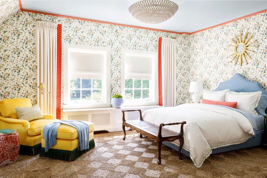 Bright bedroom with floral wallpaper, a blue upholstered bed, yellow armchair, and sunburst wall decor; brown bench at the foot of the bed