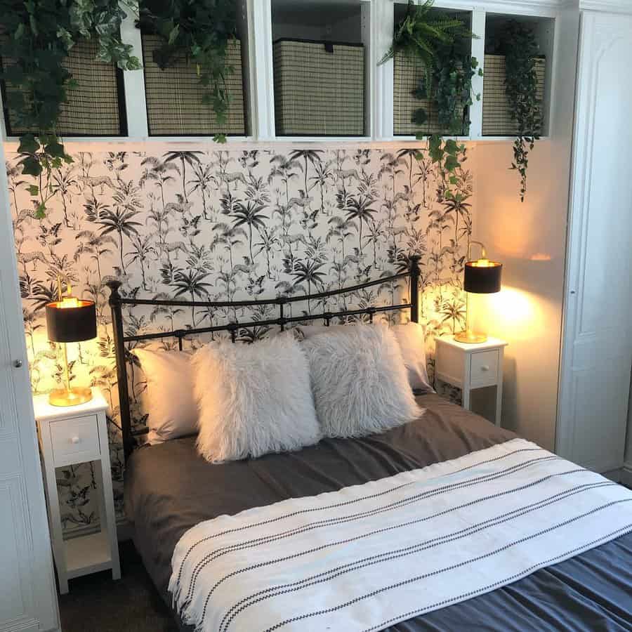 A cozy bedroom with a patterned wallpaper, metal bed frame, fluffy pillows, and hanging plants; side tables with lamps are lit