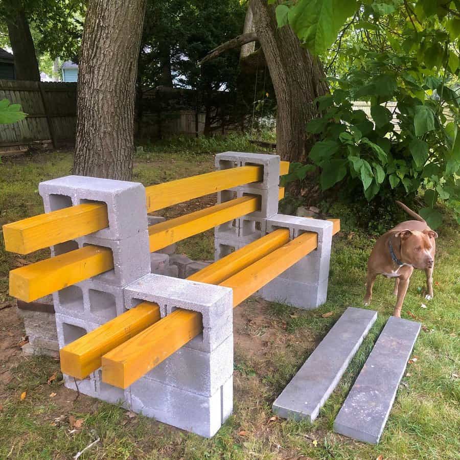 DIY outdoor bench made from cinder blocks and yellow wooden beams, set under a tree for a rustic backyard seating area