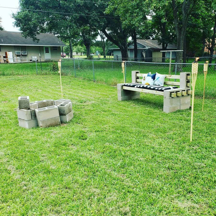 DIY backyard seating area with a cinder block bench, striped cushion, and tiki torches, set near a fire pit for a cozy outdoor gathering space