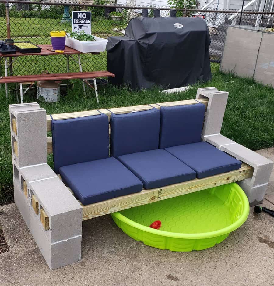 DIY cinder block bench with navy cushions, built-in armrests, and a hidden green kiddie pool underneath, creating a multi-functional outdoor space