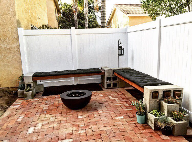 Modern outdoor seating area with L-shaped cinder block benches, black tufted cushions, a fire pit, and potted plants on a brick patio