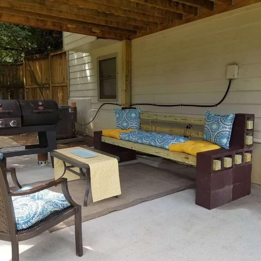 DIY cinder block bench with bright yellow and blue cushions, set under a covered patio with a grill and cozy seating area for outdoor relaxation