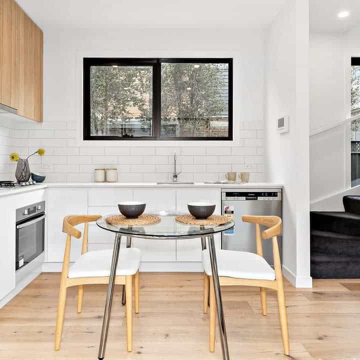 Minimalist kitchen with white cabinetry, black-framed window, round glass table, two wooden chairs, and a staircase on the right