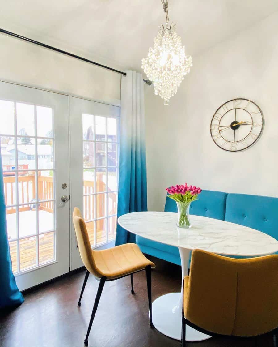 Bright dining area with a white oval table, yellow chairs, blue sofa, pink tulips in a vase, and a crystal chandelier under large windows