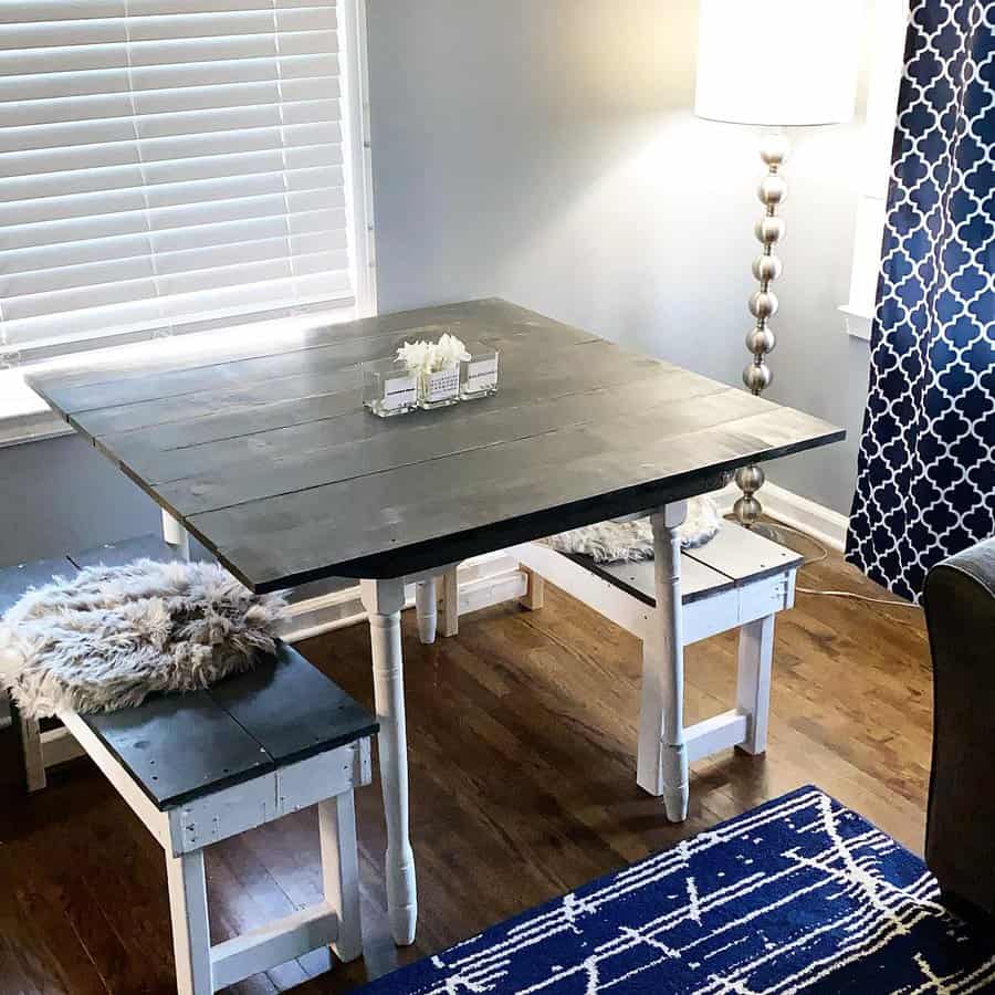 A cozy dining area with a square wooden table, two white benches with cushions, a tall lamp, and a blue patterned rug