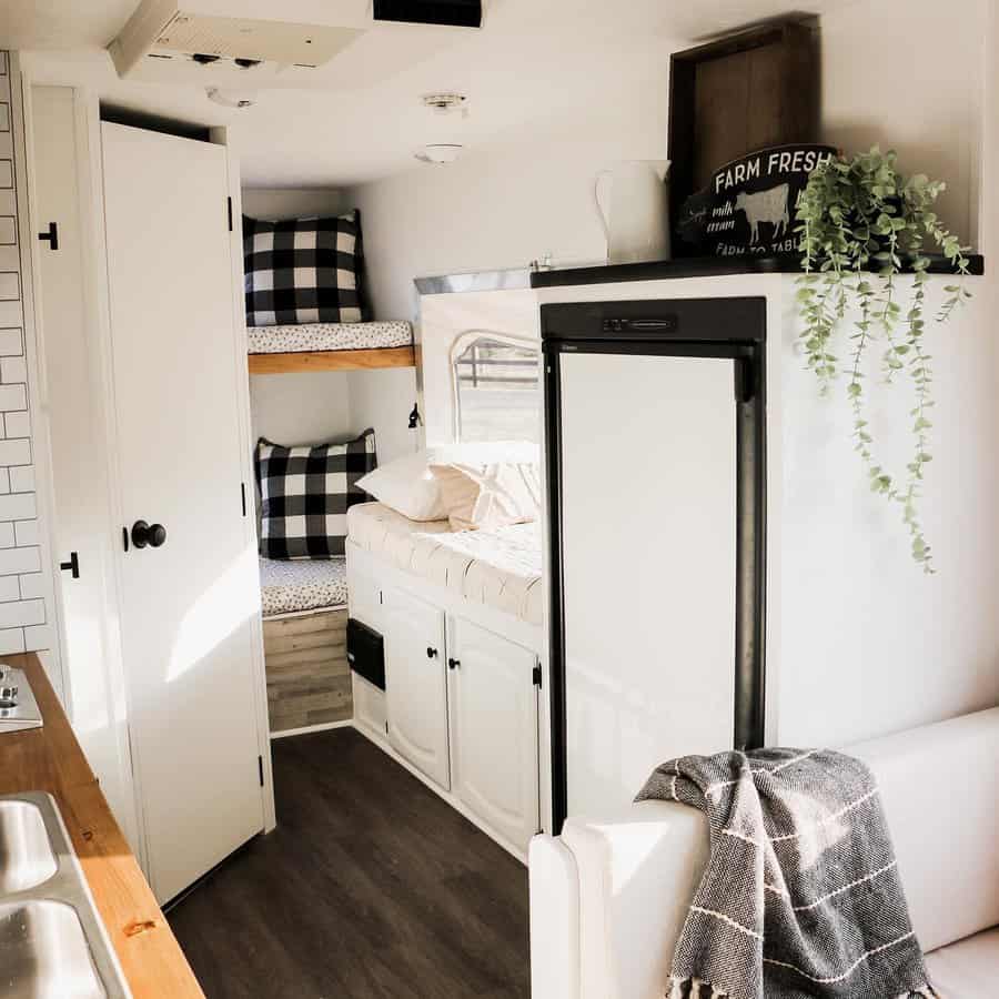 A cozy RV living space featuring bunk beds with plaid pillows, a modern fridge, a white sofa with a throw blanket, and green plants for a homely feel.