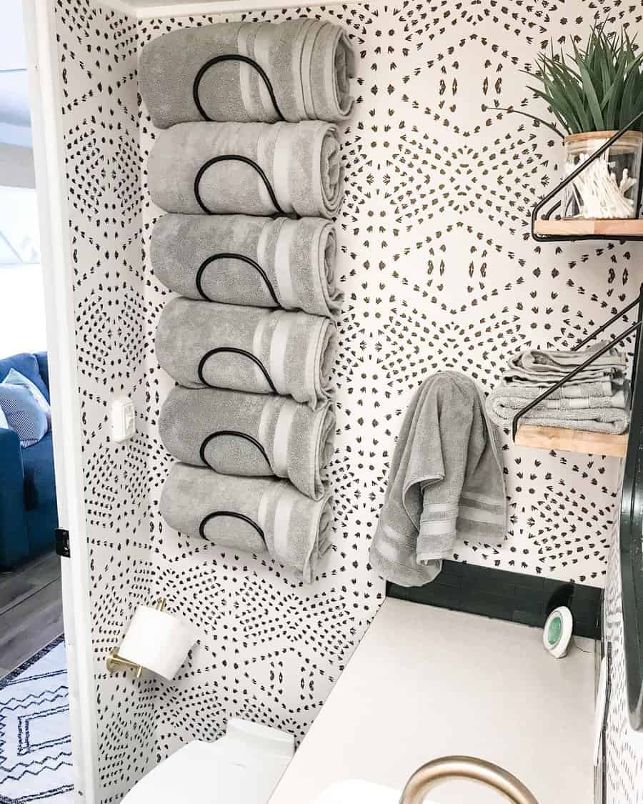 A stylish RV bathroom featuring neatly rolled gray towels on a modern towel rack, a patterned wall, and minimalistic decor with plants.