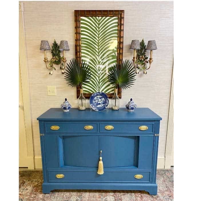 Bold blue sideboard with gold hardware, styled with palm leaves, blue and white ceramics, and a tropical mirror in an elegant entryway.