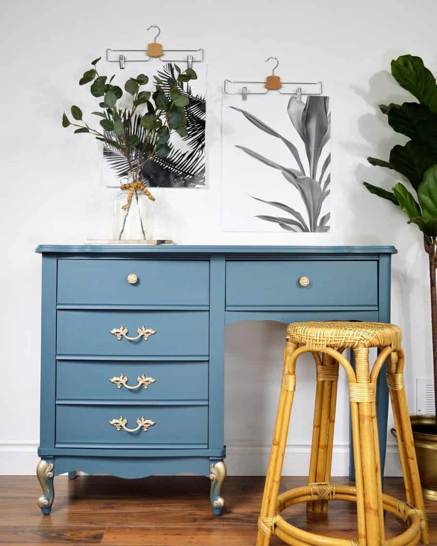 Vintage blue desk with gold hardware and curved legs, styled with a rattan stool, greenery, and monochrome botanical wall prints.