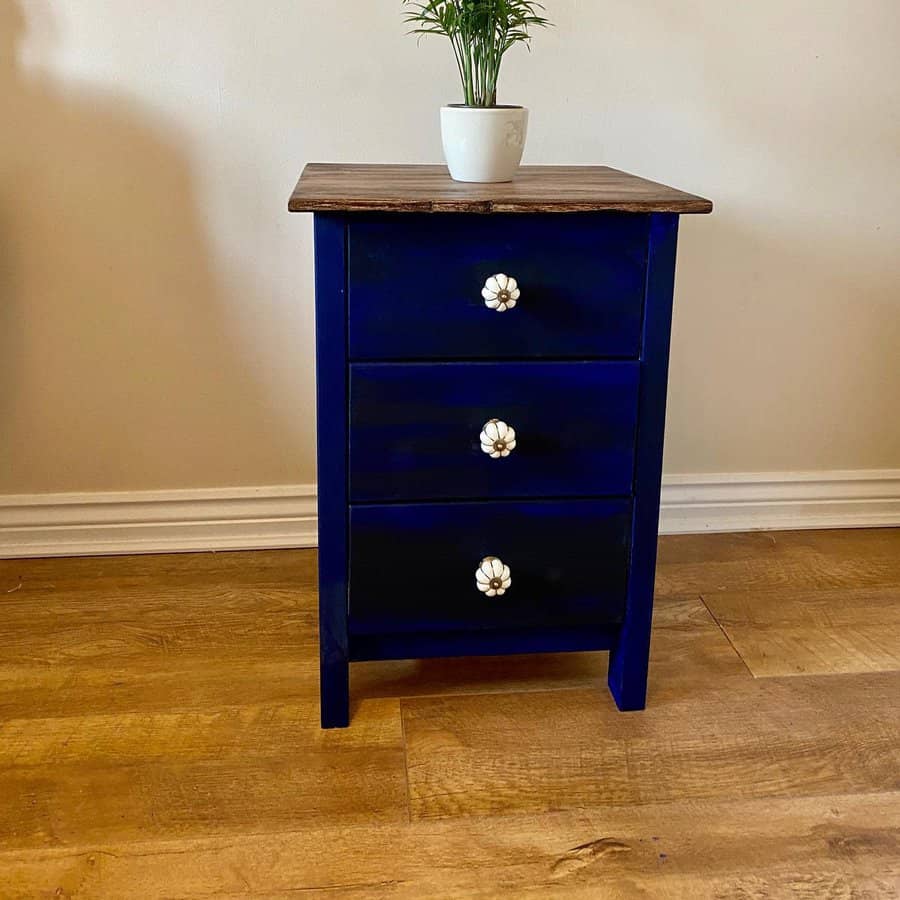 Small navy blue nightstand with a rustic wood top and white floral knobs, styled with a potted plant on a wooden floor.