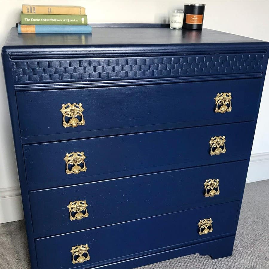 Navy blue dresser with intricate woven detailing on the top drawer and ornate gold handles, styled with books and candles on top.