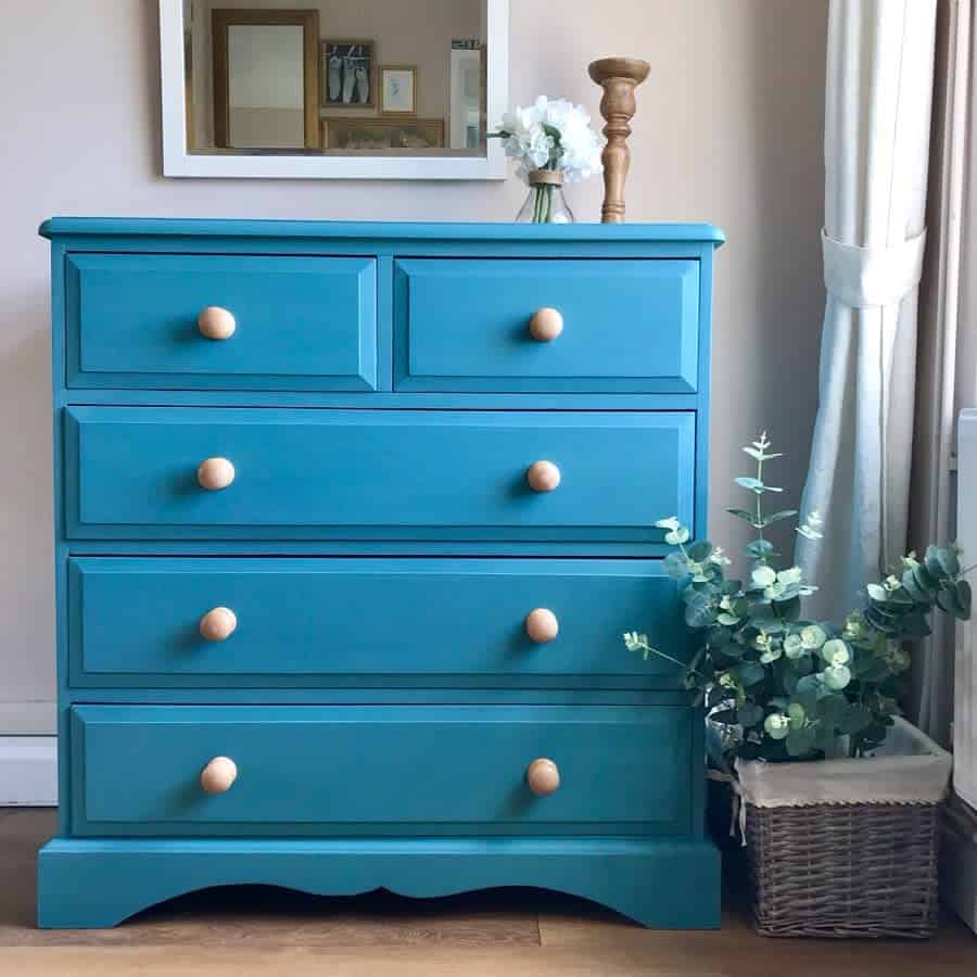 Vibrant blue dresser with natural wood knobs, styled with a mirror, floral accents, and a wicker basket filled with eucalyptus.