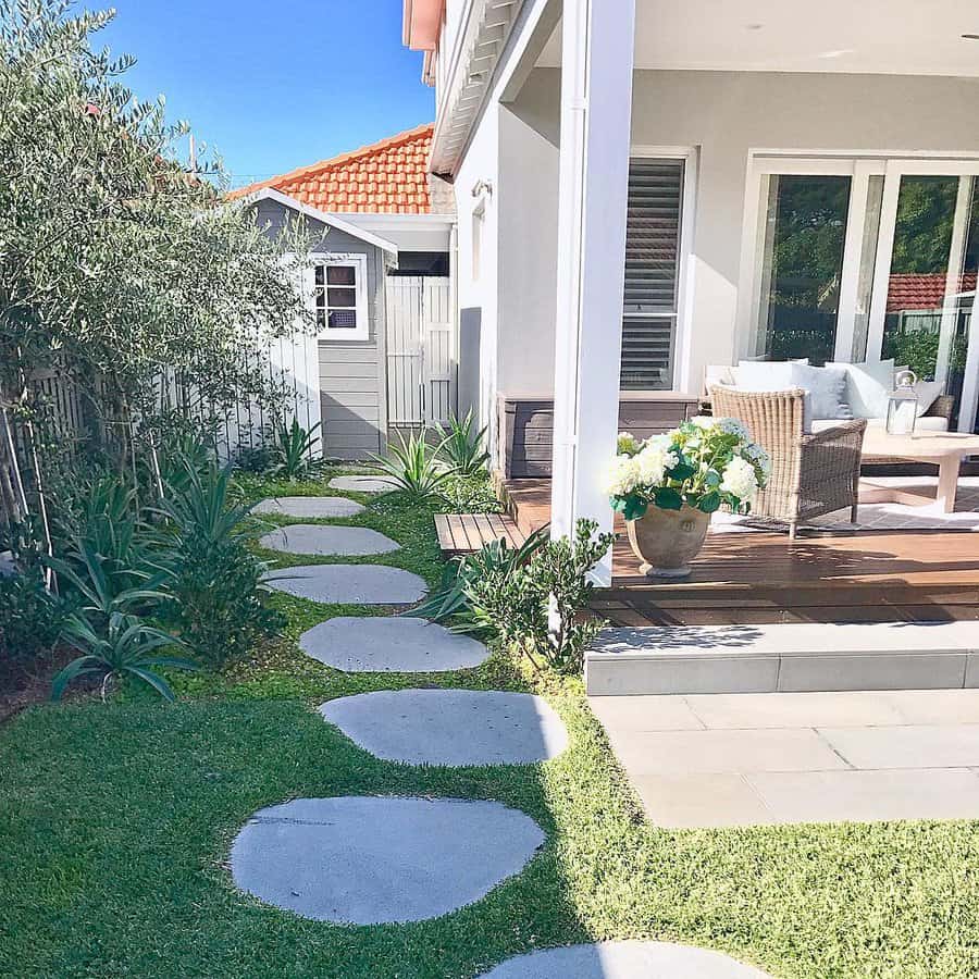 A backyard with stepping stones on grass leading to a patio featuring a wicker chair with cushions, a table, and a white shed nearby