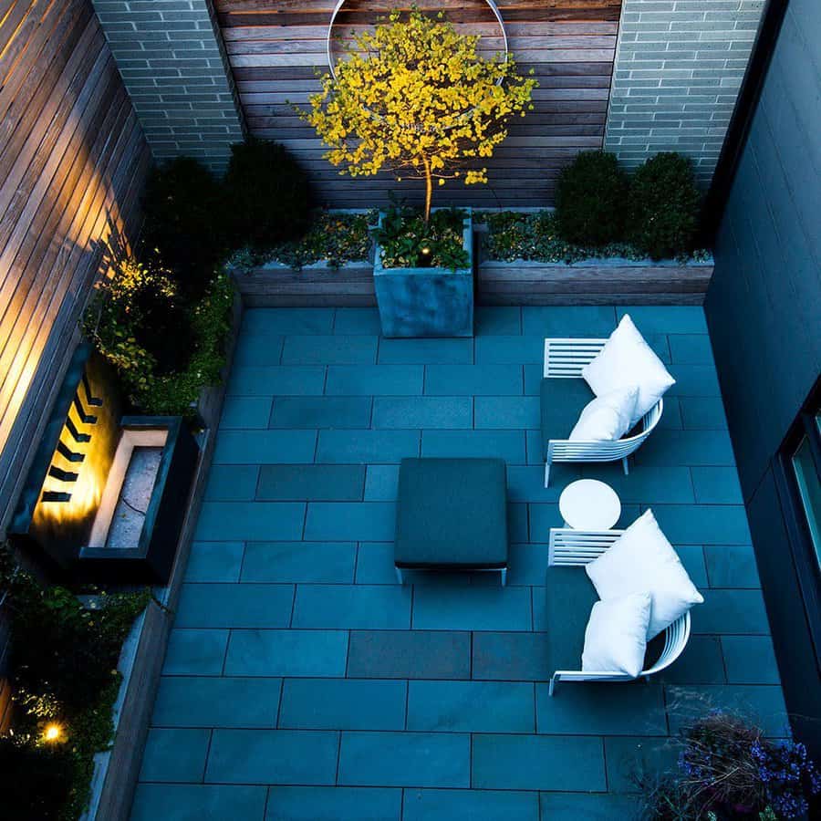 Modern backyard with sleek blue stone pavers, cozy white chairs, a fire pit, and a potted tree adding warmth and contrast