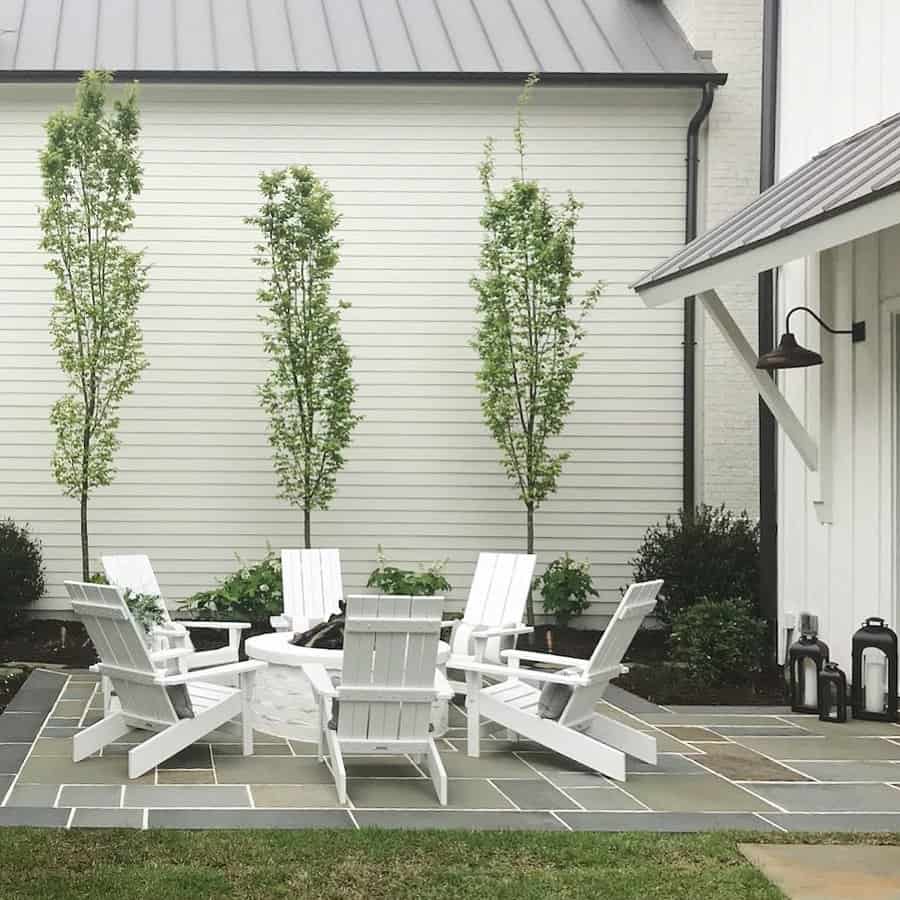 Charming backyard patio with blue stone pavers, white Adirondack chairs around a fire pit, and lush greenery for a cozy, modern farmhouse feel