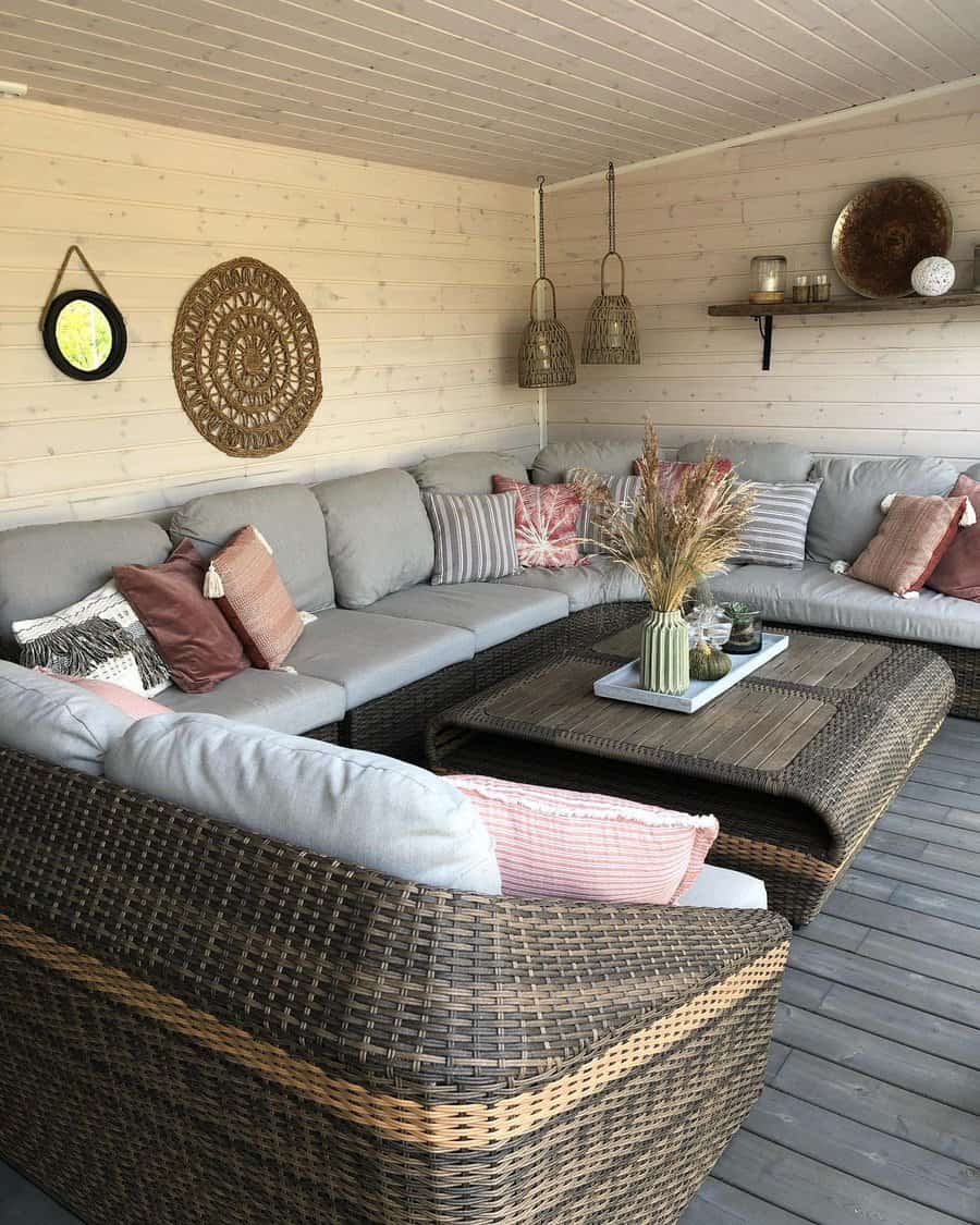 Cozy patio with L-shaped wicker sofa, gray cushions, pink pillows, decorative wall art, hanging baskets, and a coffee table with vases